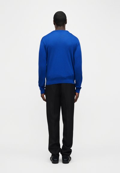 Άνδρας που στέκεται με την πλάτη, φοράει φωτεινό μπλε πουλόβερ μακρυμάνικο, μαύρο παντελόνι και μαύρα αθλητικά παπούτσια, σε λευκό απλό φόντο.