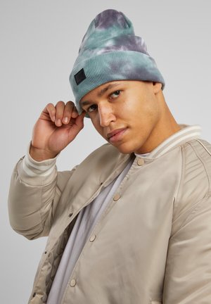 Strikket lue i teal og lilla tie-dye, med en teksturert ribbestrikket design. Personen har på seg en beige bomberjakke med beige og hvite stripete mansjetter.