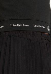 Cintura elástica negra con el logo en blanco "Calvin Klein Jeans", acompañada de un tejido plisado negro por debajo, que muestra una textura suave.