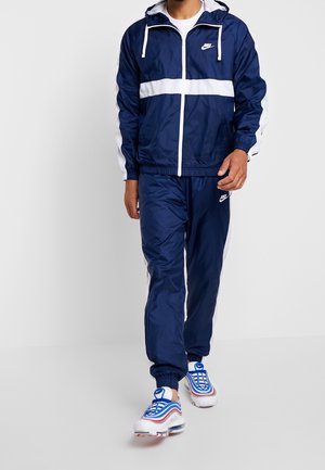 Mand iført marineblå og hvid Nike-træningsdragt med hættejakke, matchende bukser og hvide sneakers med blå og røde accenter.