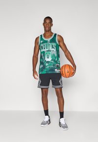 Groen basketbalshirt met de tekst "CELTICS", nummer 0 en een schubbenpatroon, gecombineerd met zwarte mesh shorts en een grijze sneaker. Houdt een basketbal vast.