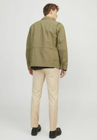 Olivgrön jacka i lätt material och avslappnad passform, parat med beiga slim-fit byxor och svarta skor, visas bakifrån.
