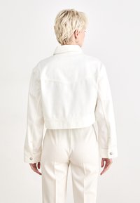 White cropped denim jacket with a collar, stitched detail on the back, and buttoned cuffs, paired with light-colored textured pants.
