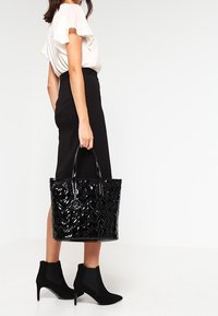 Czarna, błyszcząca torba na ramię z wytłaczanym wzorem kwiatowym, podwójnymi uchwytami i srebrnym charms. Nosi się ją z czarną spódnicą i botkami na kostkę, z falbaniastym białym topem.