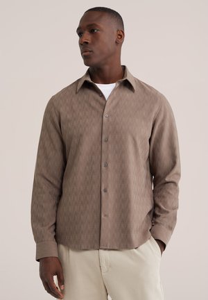 Mand iført en beige skjorte med knapper over en hvid undershirt og lyse bukser, stående med den ene hånd i lommen og kiggende mod venstre.