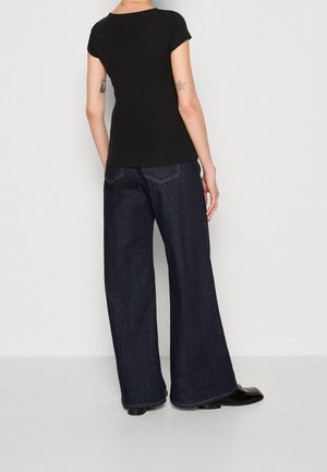 Persoon die een zwart aansluitend shirt met korte mouwen en donkere wijde jeans draagt, staat met de rug naar een eenvoudige witte muur toe.