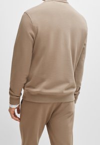 Uomo che indossa una felpa beige e pantaloni abbinati, visto da dietro su uno sfondo bianco uniforme.