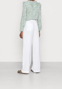 Kvinna iklädd vita vida byxor, grön blommig långärmad blus och olivgröna sandaler, stående vänd bort från kameran mot en enfärgad bakgrund.