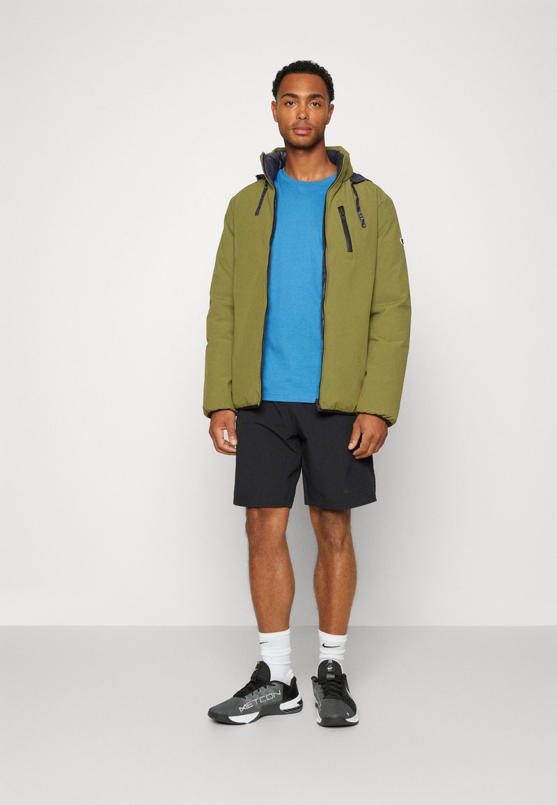 Chaqueta acolchada verde oliva con cremallera, camiseta azul, pantalones cortos negros y zapatillas negras. El modelo está de pie contra un fondo liso.