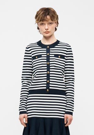 Mujer de cabello corto que lleva un cárdigan abotonado a rayas negras y blancas y una falda a juego, con un fondo liso y claro.