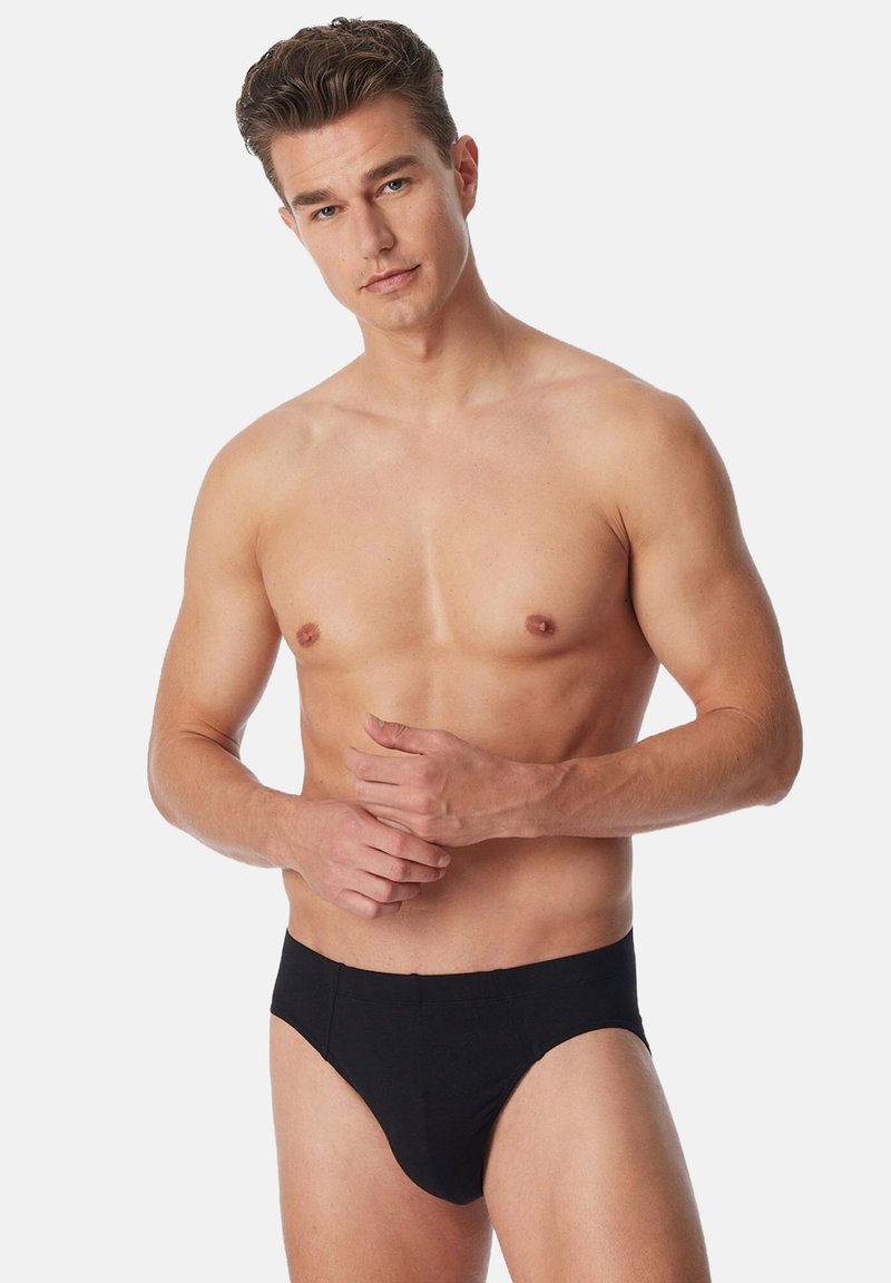 Oben ohne Mann mit kurzen braunen Haaren, der schwarze Unterwäsche trägt und mit verschränkten Händen an der Taille vor einem schlichten Hintergrund steht.