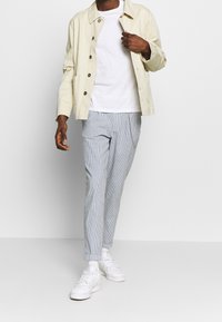 Light beige cotton jacket with buttons, white t-shirt, and striped, tapered gray pants. White sneakers and subtle chain detail on the pants.