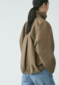 Person mit dunklen Haaren im Pferdeschwanz, die eine lockere braune Jacke mit hohem Kragen und blaue Jeans trägt, seitlich vor einem einfarbigen Hintergrund stehend.