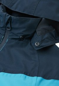 Giacca impermeabile blu navy con un accento turchese, dotata di chiusura con zip, bottoni a scatto e colletto alto con dettagli di cuciture visibili.