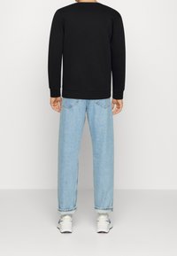 Uomo che indossa una maglietta nera a maniche lunghe, jeans arrotolati di un blu chiaro e sneakers bianche, in piedi con la schiena rivolta verso di noi su uno sfondo neutro.