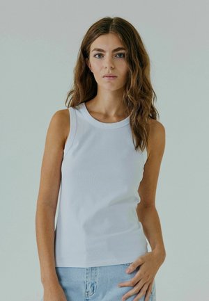 Débardeur blanc sans manches avec un col rond, fabriqué à partir d'un matériau lisse. Le vêtement épouse étroitement le corps, associé à un short en denim bleu clair.