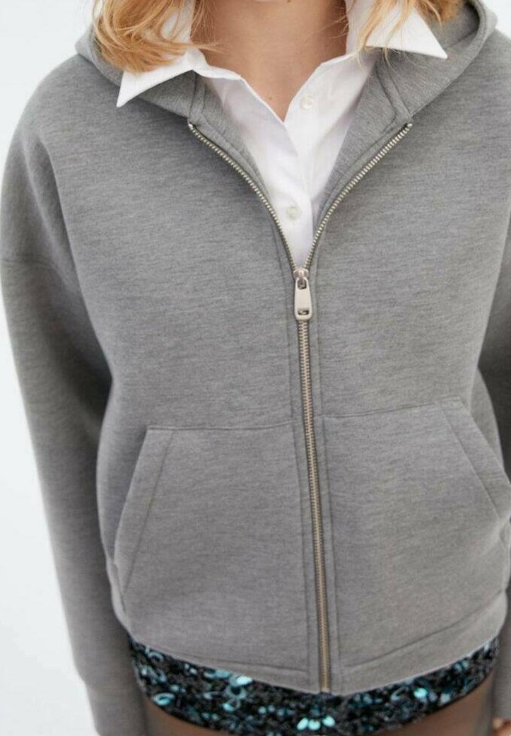 Sweat à capuche gris avec fermeture éclair, en tissu doux, équipé d'une fermeture éclair à l'avant, de deux poches latérales, et d'une capuche. Chemise blanche visible en dessous.