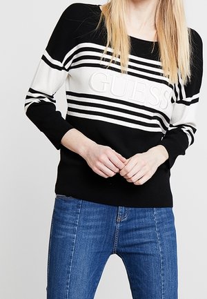 Kvinde iført sort- og hvidstribet Guess-trøje og blå jeans, stående med hænderne løst foldet foran.