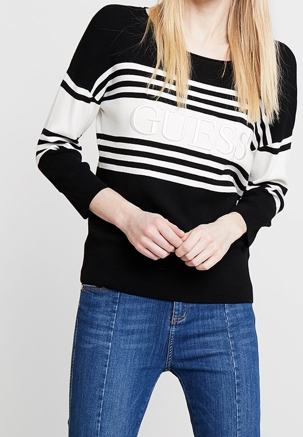 Femme portant un pull rayé noir et blanc Guess et un jean bleu, debout, les mains jointes négligemment devant elle.