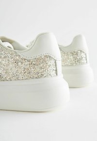 Bijele tenisice s teksturiranim gliterom na stražnjem dijelu, glatkim kožnim gornjim dijelom, okruglim prstima i gumiranim potplatom. Suptilni sjaj i minimalistički dizajn.