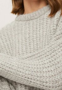 Vaaleanharmaa neulottu pusero, jossa on teksturoitu kuvio ja pyöreä pääntie, esitellen pehmeän ja kodikkaan ilmeen.