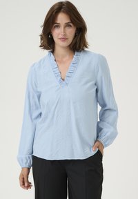 Light blue blouse with ruffled V-neckline, long sleeves, and textured fabric; paired with black pants. Simple and elegant design.