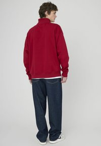 Sweatshirt rouge oversized avec un col montant, des poignets et une taille côtelés, porté avec un jean bleu foncé à coupe ample.
