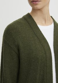 Cardigan vert forêt, tricot texturé, à l'ouverture sur le devant, avec un large col châle, porté sur une chemise blanche. Accent sur le contraste des tissus et des couleurs.