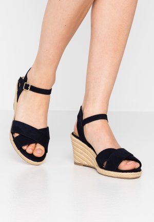 Mørkeblå kilehælssandaler med krydsede ruskindsstropper, ankelspænde og kilehæl indpakket i jute. Tæerne er synlige.