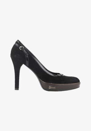 Escarpin à talon haut en daim noir avec semelle plateforme et logo Gucci sur le côté, avec un petit détail de boucle près du talon.