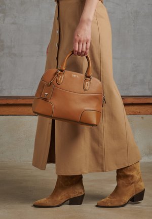 Tan leather handbag with structured shape and smooth texture, featuring dual handles and a removable tag. Worn with a long tan skirt.
