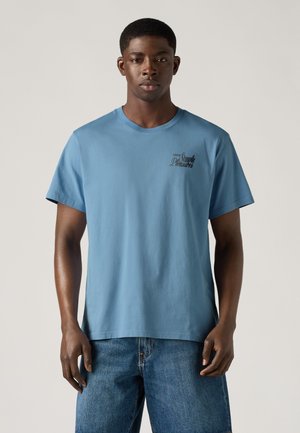 Jeune homme portant un t-shirt bleu Levi’s Simple Pleasures et un short en denim bleu, debout devant un fond clair uni.