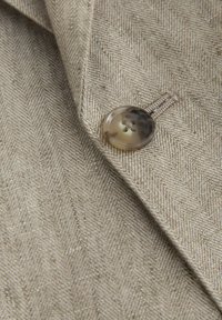 Bézs halszálka mintájú szövet részlet egy négylyukú teknőspáncélgomb közelijével, amely egy zakó hajtókájára van varrva.