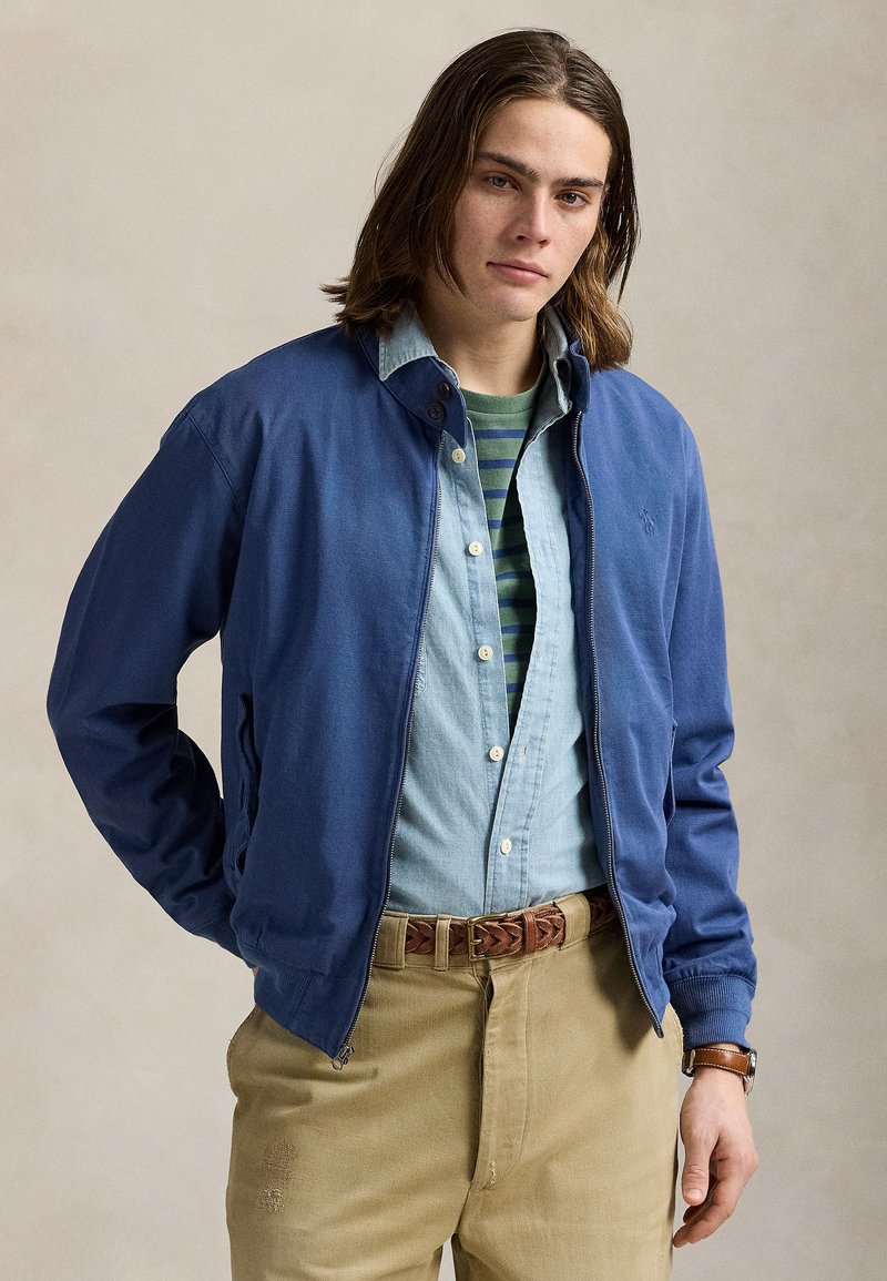 Joven con el pelo largo lleva una chaqueta azul sobre una camisa vaquera clara y una camiseta verde de rayas, pantalones beige, cinturón marrón trenzado y reloj.