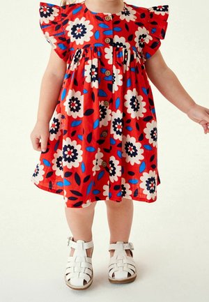 Tout-petit portant une robe rouge avec un grand imprimé floral blanc, bleu et noir, et des sandales blanches à boucles, devant un fond uni.