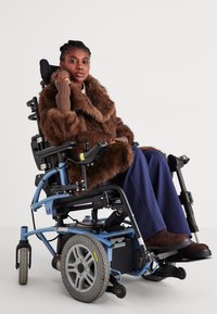Γυναίκα με καφέ γούνινο παλτό καθισμένη σε ένα μπλε ηλεκτρικό αναπηρικό καροτσάκι, με φόντο λευκό.