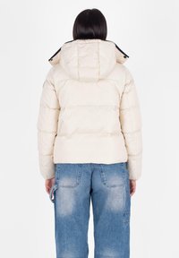 Puchowa beżowa kurtka z kapturem, charakteryzująca się gładką powierzchnią i pikowaniem. W połączeniu z luźnymi niebieskimi dżinsami, które eksponują szczegóły tylnej kieszeni.