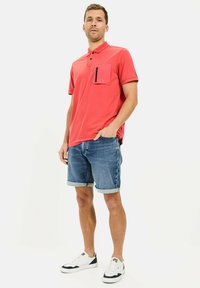 Rotes Poloshirt mit einer Brusttasche, kombiniert mit hochgekrempelten Jeansshorts und weißen Sneakers mit navyfarbenen Akzenten. Minimalistisches Design.