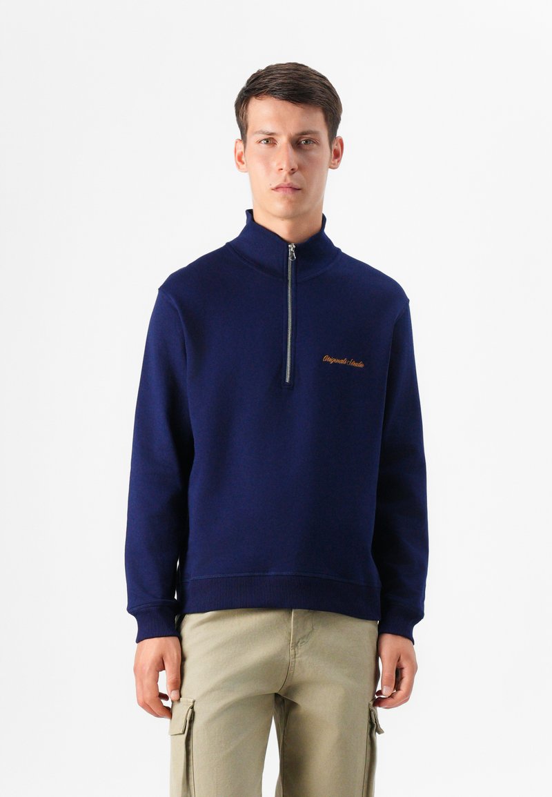 Un giovane uomo si trova di fronte a uno sfondo bianco semplice, indossando una felpa con zip blu navy e pantaloni cargo di colore chiaro.