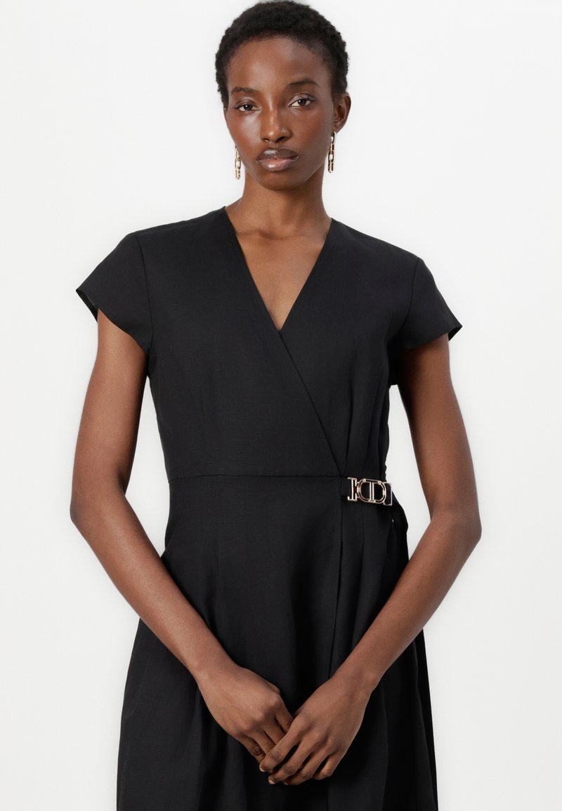 Czarna sukienka kopertowa z krótkimi rękawami, z dekoltem w kształcie litery V i metalowym zapięciem w pasie. Gładka faktura materiału.