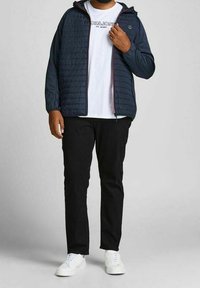 Mörkblå quiltad jacka med huva, bärs över en vit t-shirt, kombineras med svarta byxor och vita sneakers. Har dragkedjestängning och synlig logga.