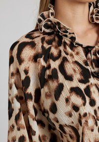 Blusa de estampado de leopardo con textura plisada, cuello negro con volantes y base beige con manchas marrones oscuras y negras.