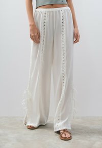 Pantalones blancos fluidos con inserciones de encaje a lo largo de los laterales, con lazos en los dobladillos. Hechos de una tela ligera y con un corte relajado.