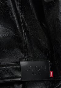 Schwarzer Kunstlederstoff mit strukturiertem Finish. Verfügt über ein rechteckiges schwarzes Etikett mit der Prägung "JOOP! JEANS" und einem roten Akzentetikett.