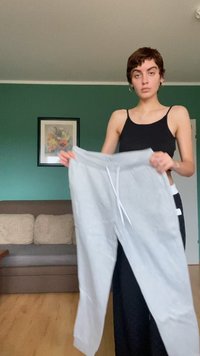 Ljusgrå joggingsbyxor med elastisk midja, dragsko och mudd vid anklarna. Tillverkade av mjukt tyg, utställda mot en grön vägg.