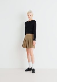 Haut noir à manches longues, jupe plissée beige et chaussures noires avec des chaussettes blanches. Le mannequin se tient devant un fond blanc uni.