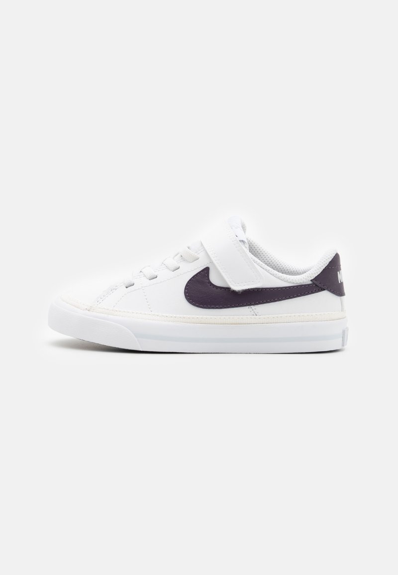Baskets basses blanches avec le logo Nike noir, lacets et une bride Velcro sur un fond clair uni, vue de côté face à gauche.