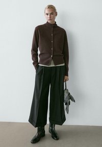 Bruine cardigan met een opstaande kraag en knopen, gecombineerd met een grijze top en brede zwarte broek. Zwarte laarzen en een accessoires met patroon in de hand.