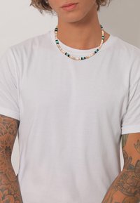 Hvid crew-neck t-shirt båret med en flerfarvet perlekæde, der har beige, blå og sorte perler, hvilket fremhæver en glat tekstur.