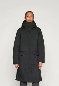 Woman wearing a long black winter coat with high collar and front flap pockets, standing against a light gray background.